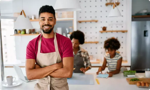 A man wearing an apron stands with arms crossed in a kitchen. In the background, a woman and a child are working at a counter.