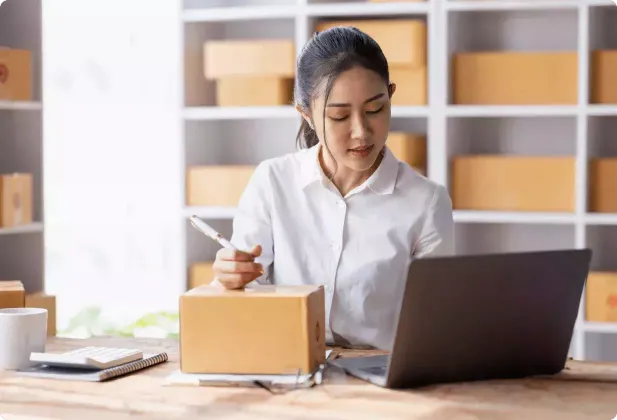 A woman in a white shirt works at a desk with a laptop, holding a pen and looking at a small cardboard box. Shelves with more boxes are in the background.